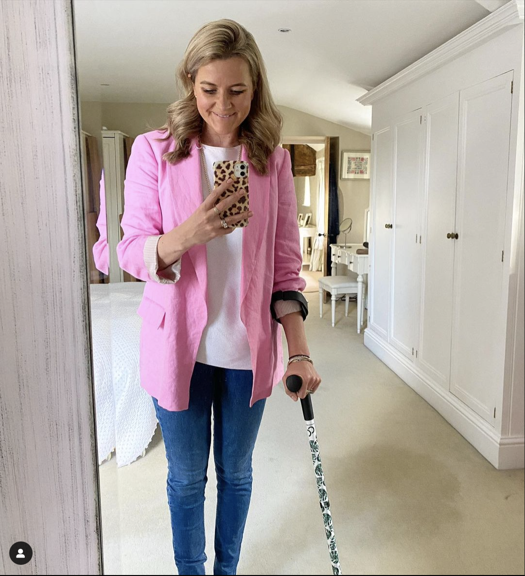 Younger middle aged white woman with shoulder length blonde hair tucked behind her ears. She's smiling while looking at her animal print covered phone held in her right hand which is the left side of the photo. She is wearing a bright pink blazer and blue jeans. She is using a forearm crutch with her left hand and arm. The crutch has black forearm cuff and handle. The stick part is white with a green leaf design.