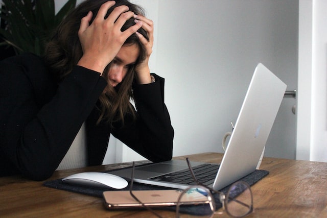 Woman with long dark hair holding her head seemingly in frustration over a silver laptop computer on a white desk.