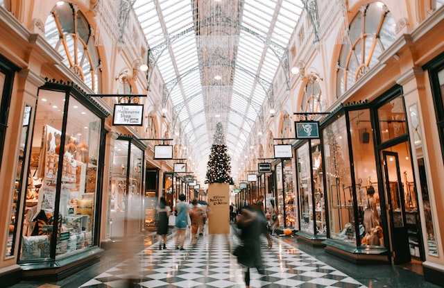 Inside a shopping mall looking down an aisle of shops. The stores are decorated for Christmas. There is a Christmas tree on a raised stand in the middle of the photo.