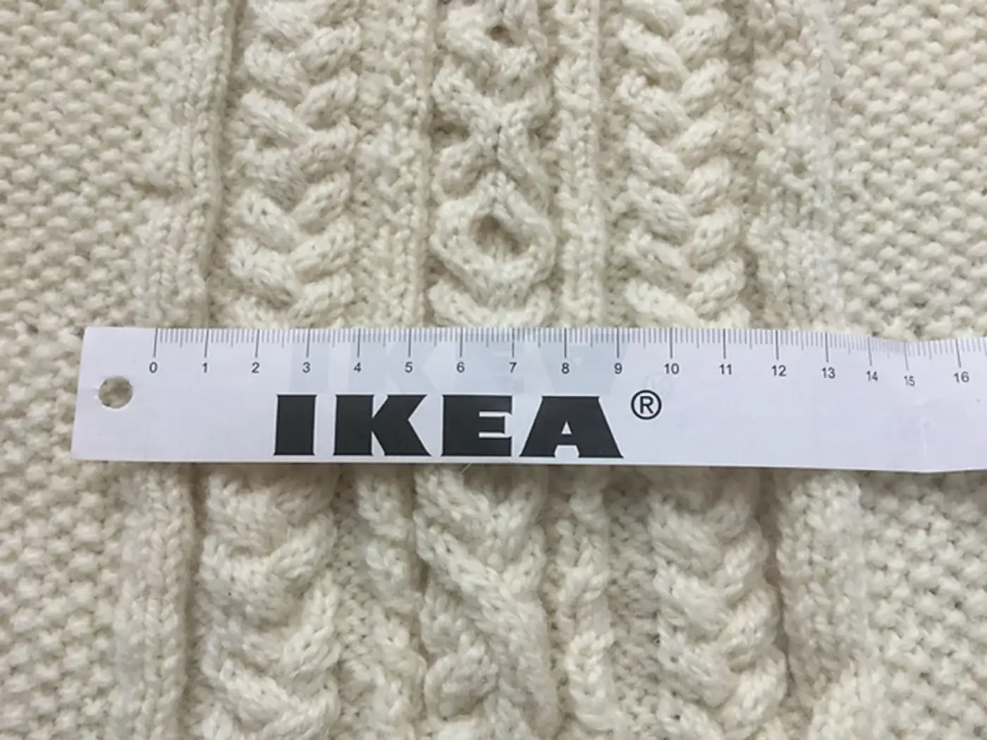 Closeup of a light cream knitted cardigan cable panel detail. There's a paper Ikea measuring tape to provide scale. The panel is about 13 cm wide.