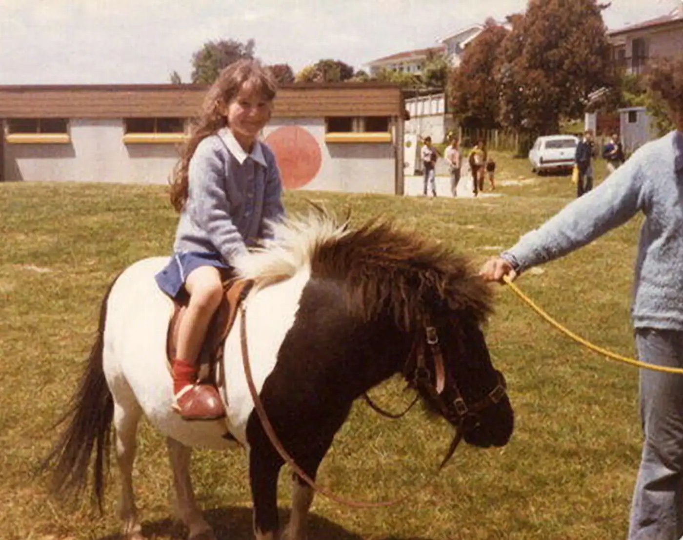 The author riding a pony at Discovery School in Whitby, Wellington Region, New Zealand. She appears about 9 years old with long wavy hair. She is wearing a light blue cardigan, a skirt, and socks. A woman is leading the pony